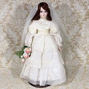 May include: A porcelain doll dressed as a bride, wearing a white wedding gown with a layered skirt and long sleeves. The doll has brown hair, a veil, and holds a bouquet of pink and white flowers. The doll is standing on a dark brown base.