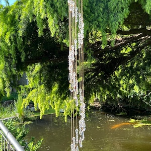 Pode incluir: Um carrilhão decorativo com contas de cristal transparente em forma de gota penduradas em correntes douradas. O carrilhão está suspenso de um galho de árvore, com um lago e vegetação ao fundo. Os cristais capturam a luz do sol.