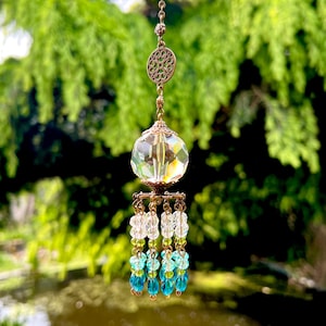 May include: A crystal suncatcher with a clear crystal ball and a decorative metal top. The suncatcher has a chain with a decorative metal charm. The crystal ball is surrounded by strands of clear, light green, and teal beads.