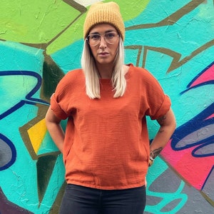 Könnte beinhalten: Eine Frau trägt ein rostfarbenes, kurzärmeliges Hemd, schwarze Jeans und weiße Turnschuhe. Sie steht vor einer bunten Graffitiwand und trägt eine gelbe Mütze.