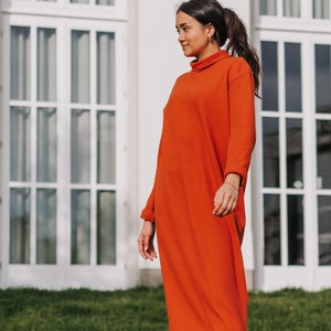 Könnte beinhalten: Eine Frau trägt ein langes, rostfarbenes Rollkragenkleid mit gerippter Struktur. Sie steht vor einem weißen Gebäude mit großen Fenstern.