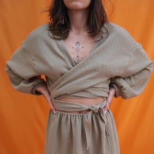 Könnte beinhalten: Zweiteiliges Outfit in hellem Olivgrün. Das Oberteil ist eine Wickelbluse mit langen Ärmeln. Der Rock ist ein Minirock mit Kordelzug in der Taille. Der Stoff hat eine strukturierte, leicht zerknitterte Optik. Der Hintergrund ist einfarbig orange.