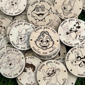 May include: A collection of round wooden tokens with engraved cartoon character illustrations. Each token features a different character and the text "Thank You For Making The Magic" in a circular layout.