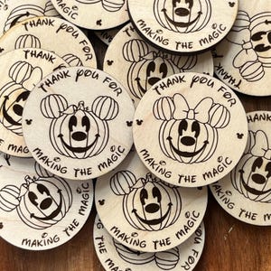 May include: A collection of round wooden coasters featuring a pumpkin-Mickey Mouse hybrid design. The coasters have the phrases "Thank You For" and "Making the Magic" etched around the edges. Some have a bow.