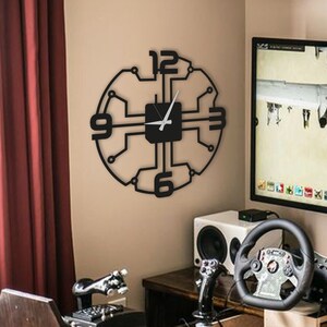 May include: A black clock with a circuit board design and silver hands. The clock is mounted on a wall in a room with a gaming setup, including a steering wheel, pedals, and a computer monitor.