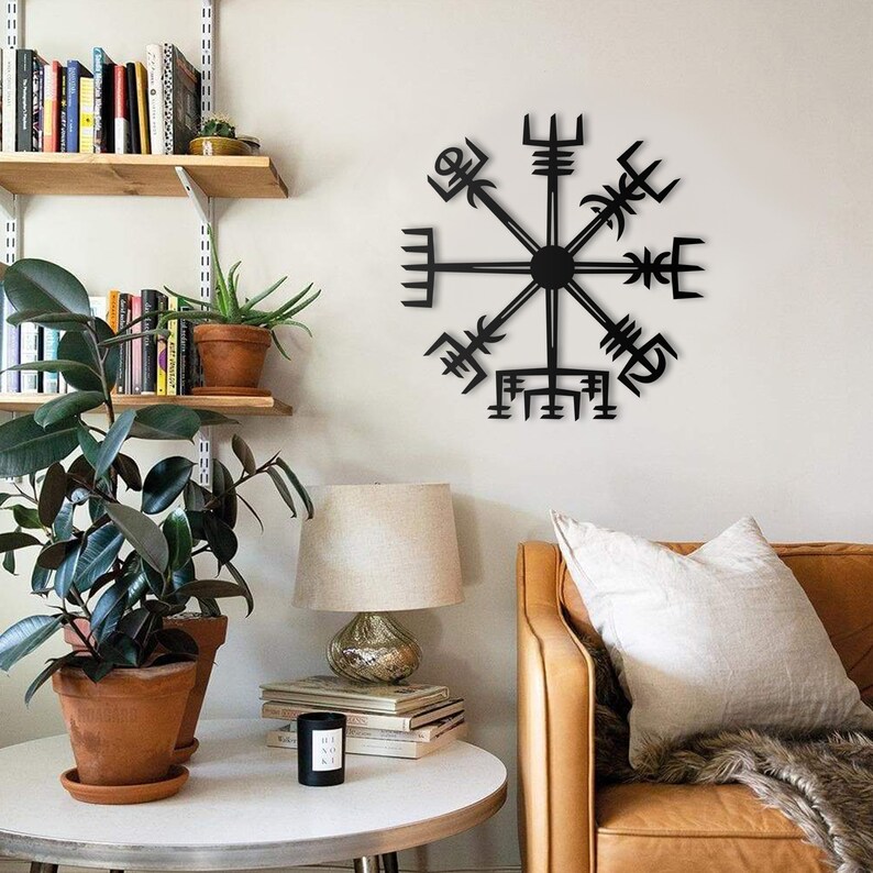 May include: A black metal Vegvisir symbol, a Viking compass, is mounted on a white wall. The symbol has eight arms radiating from a central circle. The room features a leather sofa, a side table with a lamp, and potted plants, creating a cozy, decorative space.