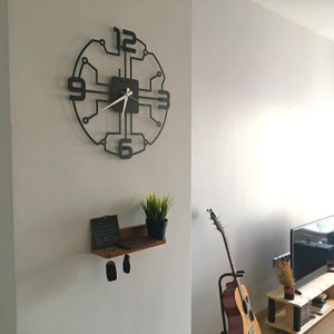 May include: A black metal clock with a circuit board design and white hands. The clock is mounted on a white wall above a wooden shelf with a small potted plant, a brown leather wallet, and a black card.