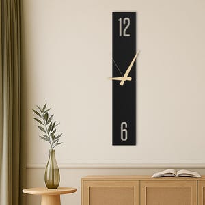 May include: A modern, rectangular wall clock with a black face. The clock features silver numerals at the 12 and 6 positions, with light-colored wooden hands. The clock is mounted on a light-colored wall, adding a minimalist touch to the decor.