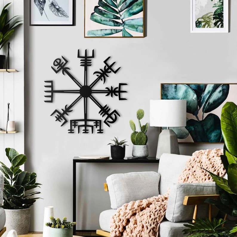 May include: A black metal wall art piece featuring the Vegv&iacute;sir symbol, a Viking compass. The artwork is mounted on a white wall, surrounded by framed botanical prints and indoor plants, creating a modern, minimalist aesthetic.