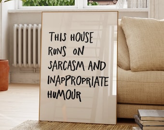 May include: A rectangular framed wall art print featuring a light beige background with black hand-lettered text that reads: This house runs on sarcasm and inappropriate humour. The print is displayed standing on a light-colored wooden floor.