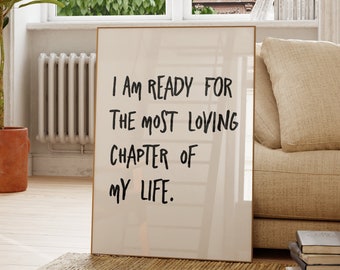 May include: A rectangular framed wall art piece featuring a white background with black handwritten-style text that reads: I AM READY FOR THE MOST LOVING CHAPTER OF MY LIFE. The frame is thin and light-colored, standing upright on a woven rug.