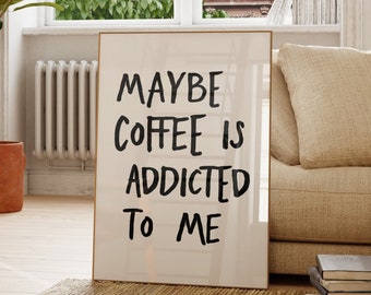 May include: A rectangular framed wall art piece featuring a white background with black hand-lettered text that reads: MAYBE COFFEE IS ADDICTED TO ME. The frame is thin and light-coloured, positioned upright on a woven rug in a bright room.