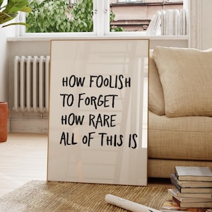 May include: A framed art print with the text "HOW FOOLISH TO FORGET HOW RARE ALL OF THIS IS" leans against a beige sofa. A potted plant and wooden chair are in the foreground. The room has a neutral color palette with natural light.