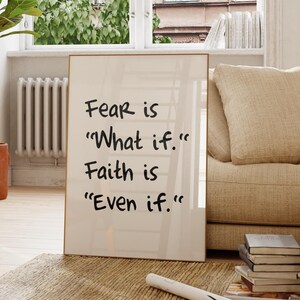 Peut inclure: Un cadre avec le texte "Fear is 'What if.' Faith is 'Even if.'" est posé sur un tapis dans un salon. La pièce comprend une chaise en bois, une plante en pot et un canapé beige. Une fenêtre offre une vue sur un bâtiment.