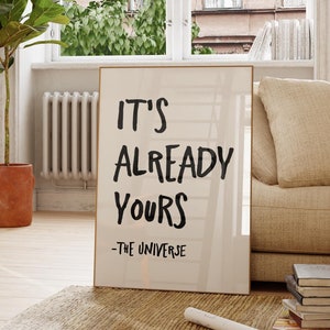Pode incluir: Uma impressão emoldurada com o texto "It's Already Yours - The Universe" em escrita preta sobre fundo branco. A impressão está encostada em um sofá bege em uma sala de estar com uma janela grande e uma planta em vaso.