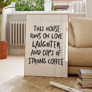 Op de afbeelding: Een ingelijste print met de tekst "THIS HOUSE RUNS ON LOVE LAUGHTER AND CUPS OF STRONG COFFEE" leunt tegen een beige bank. Een plant in een pot en een houten stoel staan op de voorgrond. De kamer heeft een neutraal kleurenpalet.