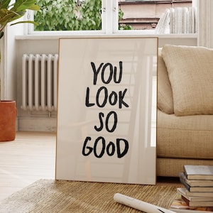 以下が含まれることがあります： 白い背景に太字の手書き風スタイルで「YOU LOOK SO GOOD」という黒いテキストが印刷された、長方形の額入りウォールアート。フレームは細い金属製で、明るい部屋の織りラグの上に立てかけられています。