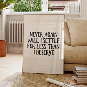 Può includere: Un poster con una citazione motivazionale con il testo "Never again will I settle for less than I deserve" in lettere nere su sfondo bianco. Il poster è incorniciato da una cornice dorata sottile e appoggia su un divano beige in un soggiorno.