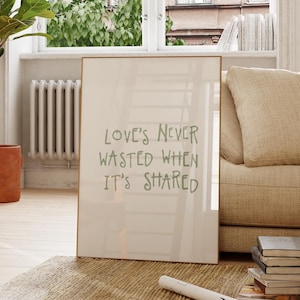 Könnte beinhalten: Ein rechteckiges gerahmtes Wandbild mit hellbeigem Hintergrund und dem dunkelgrünen Text "LOVE'S NEVER WASTED WHEN IT'S SHARED" in einer einfachen, handgezeichneten Schriftart, das aufrecht auf einem Webteppich steht.