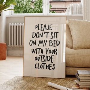 Pode incluir: Uma peça de arte retangular emoldurada com fundo branco e texto preto manuscrito em negrito que diz: PLEASE DON'T SIT ON MY BED WITH YOUR OUTSIDE CLOTHES. A moldura é de madeira fina de cor clara e está apoiada no chão.