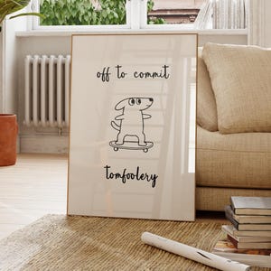 May include: A framed print featuring the text "off to commit tomfoolery" and a cartoon dog on a skateboard. The print is propped against a beige sofa in a bright room with a large window and a potted plant.