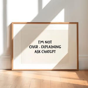 May include: A framed print with the text "I'M NOT OVER - EXPLAINING ASK CHATGPT" in black font. The print is in a room with hardwood floors and a white wall. The frame is a light brown color.