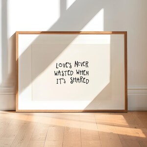 May include: A rectangular framed print with a light beige background and black hand-lettered text that reads, "LOVE'S NEVER WASTED WHEN IT'S SHARED." The frame is made of light-coloured wood and features a clean, simple border design.
