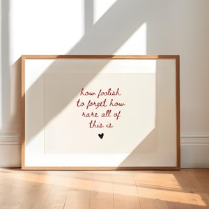 May include: A framed print with the quote "how foolish to forget how rare all of this is" in red script, with a small black heart. The frame is a light brown color, and the print is displayed on a wooden floor.