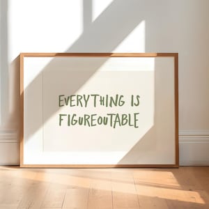 Op de afbeelding: Een ingelijste print met de tekst "EVERYTHING IS FIGUREOUTABLE" in groene letters. De print zit in een lichtgekleurde houten lijst en ligt op een houten vloer. De kamer heeft witte muren en natuurlijk licht.