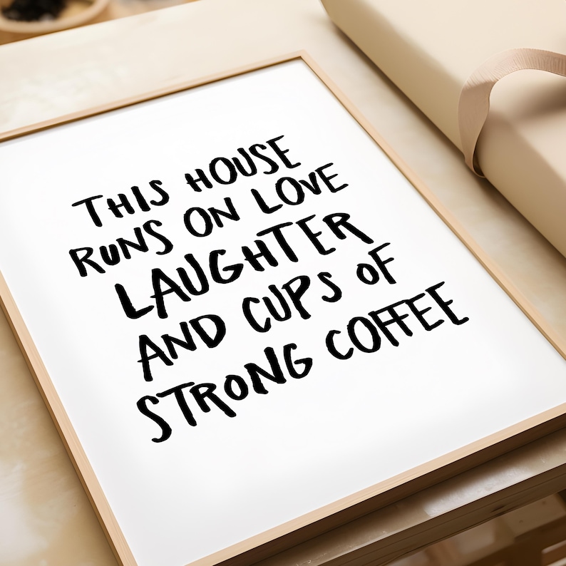 Op de afbeelding: Een ingelijste print met de tekst "THIS HOUSE RUNS ON LOVE LAUGHTER AND CUPS OF STRONG COFFEE" in zwart op een witte achtergrond. De lijst is licht van kleur. Een beige geschenkdoos is gedeeltelijk zichtbaar.