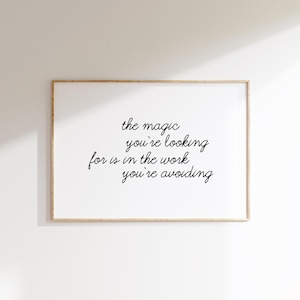 May include: A rectangular wall art print in a light wood frame featuring the black cursive text: the magic you're looking for is in the work you're avoiding. The print is centred on a plain white background with a soft shadow cast across the top.