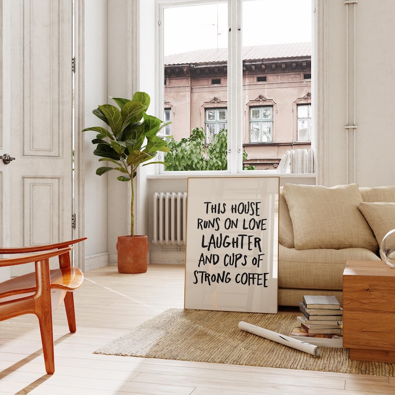 Op de afbeelding: Een ingelijste print met de tekst "THIS HOUSE RUNS ON LOVE LAUGHTER AND CUPS OF STRONG COFFEE" leunt tegen een beige bank. Een plant in een pot en een houten stoel staan op de voorgrond. De kamer heeft een neutraal kleurenpalet.