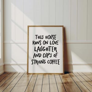 Op de afbeelding: Een ingelijste print met de tekst "THIS HOUSE RUNS ON LOVE LAUGHTER AND CUPS OF STRONG COFFEE" in zwarte letters. De lijst is lichtbruin en de achtergrond is wit. De print wordt weergegeven op een houten vloer.