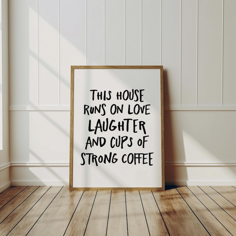 Op de afbeelding: Een ingelijste print met de tekst "THIS HOUSE RUNS ON LOVE LAUGHTER AND CUPS OF STRONG COFFEE" in zwarte letters. De lijst is lichtbruin en de achtergrond is wit. De print wordt weergegeven op een houten vloer.