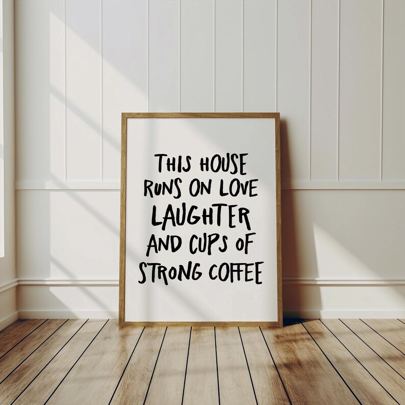 Op de afbeelding: Een ingelijste print met de tekst "THIS HOUSE RUNS ON LOVE LAUGHTER AND CUPS OF STRONG COFFEE" in zwarte letters. De lijst is lichtbruin en de achtergrond is wit. De print wordt weergegeven op een houten vloer.