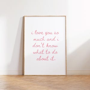 May include: A framed print with the text "i love you so much and i don't know what to do about it" in pink script. The print is in a light wood frame and is displayed against a white wall and a wooden floor.