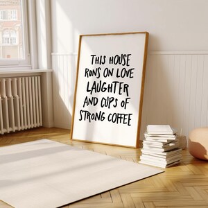 Op de afbeelding: Een ingelijste print met de tekst "THIS HOUSE RUNS ON LOVE LAUGHTER AND CUPS OF STRONG COFFEE" in zwarte letters. De lijst is lichtbruin van kleur. De print leunt tegen een witte muur. Een stapel boeken staat ernaast.