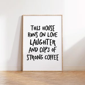 Op de afbeelding: Een ingelijste print met de tekst "THIS HOUSE RUNS ON LOVE LAUGHTER AND CUPS OF STRONG COFFEE" in zwarte letters. De lijst is van licht hout en de print staat op een houten vloer.