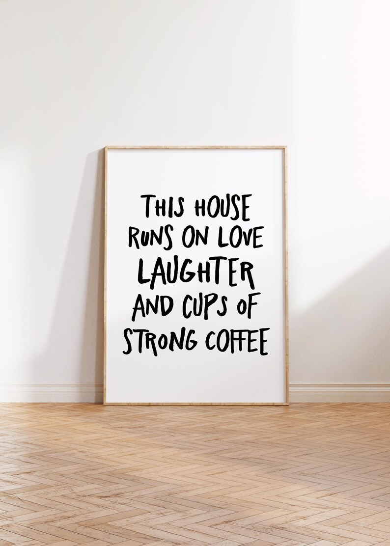 Op de afbeelding: Een ingelijste print met de tekst "THIS HOUSE RUNS ON LOVE LAUGHTER AND CUPS OF STRONG COFFEE" in zwarte letters. De lijst is van licht hout en de print staat op een houten vloer.