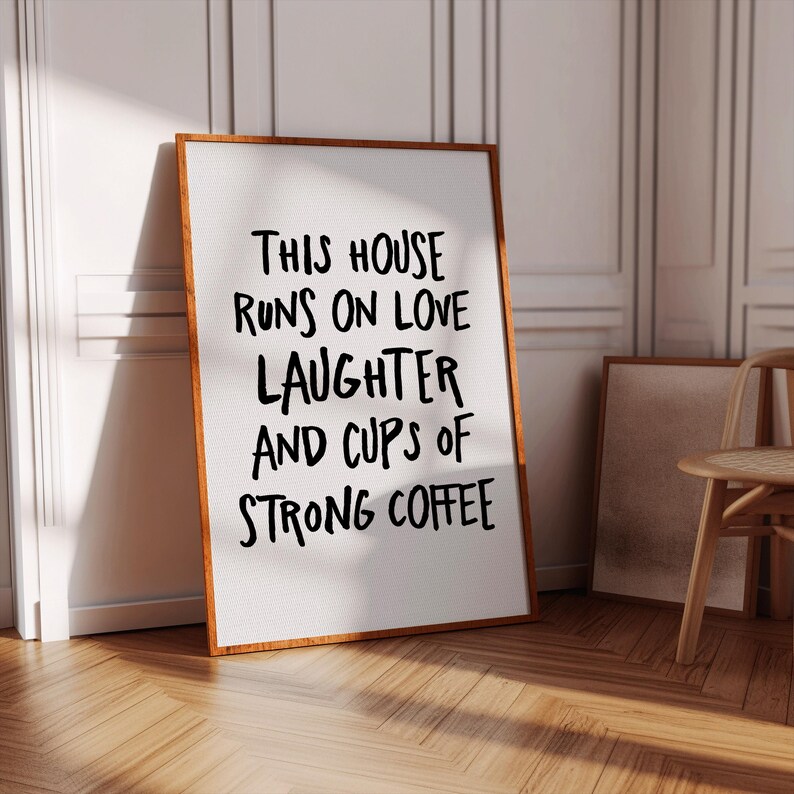 Op de afbeelding: Een ingelijste print met de tekst "THIS HOUSE RUNS ON LOVE LAUGHTER AND CUPS OF STRONG COFFEE" in zwarte letters. De lijst heeft een warme bruine kleur en de print leunt tegen een witte muur.