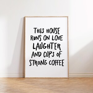Op de afbeelding: Een ingelijste print met de tekst "THIS HOUSE RUNS ON LOVE LAUGHTER AND CUPS OF STRONG COFFEE" in zwarte letters. De print zit in een licht houten lijst en wordt weergegeven tegen een witte muur en een houten vloer.