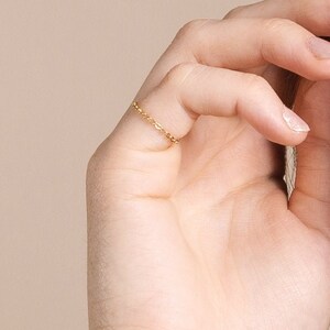 May include: A delicate gold ring with a chain-like design is worn on a finger. The image is a close-up, showing the ring's intricate details and the skin's texture. The background is a soft, neutral color, highlighting the jewelry.