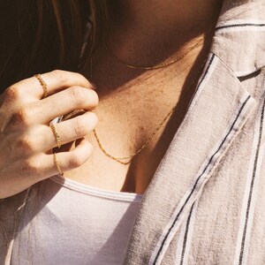 May include: Close-up of gold jewelry, including a delicate chain necklace and several chain rings. The jewelry is worn with a white top and a striped blazer. The image has a warm, natural lighting.