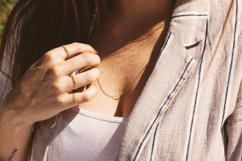 May include: Close-up of gold jewelry, including a delicate chain necklace and several chain rings. The jewelry is worn with a white top and a striped blazer. The image has a warm, natural lighting.