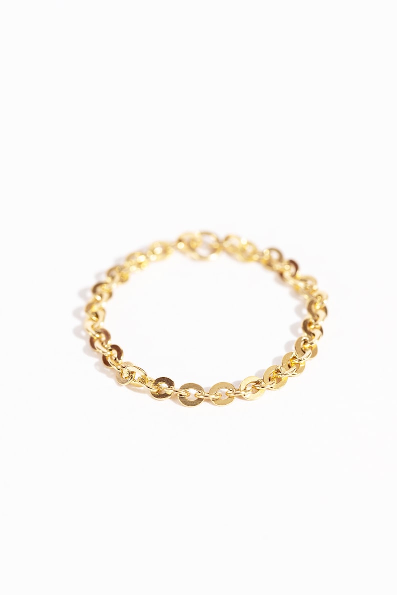 May include: A delicate gold bracelet with a repeating pattern of small, flat, circular links. The bracelet is displayed on a white background, highlighting its shiny, reflective surface. The jewelry piece is a simple yet elegant accessory.