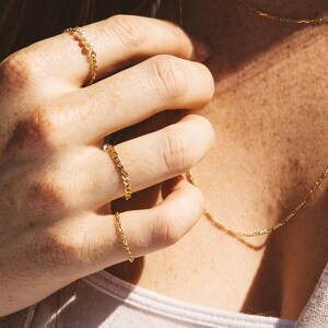 May include: A close-up shot of gold jewelry, including a chain necklace and three rings. One ring features a delicate chain design with three small pearls. The other two rings have a curb chain design. The jewelry is worn on a person's hand.