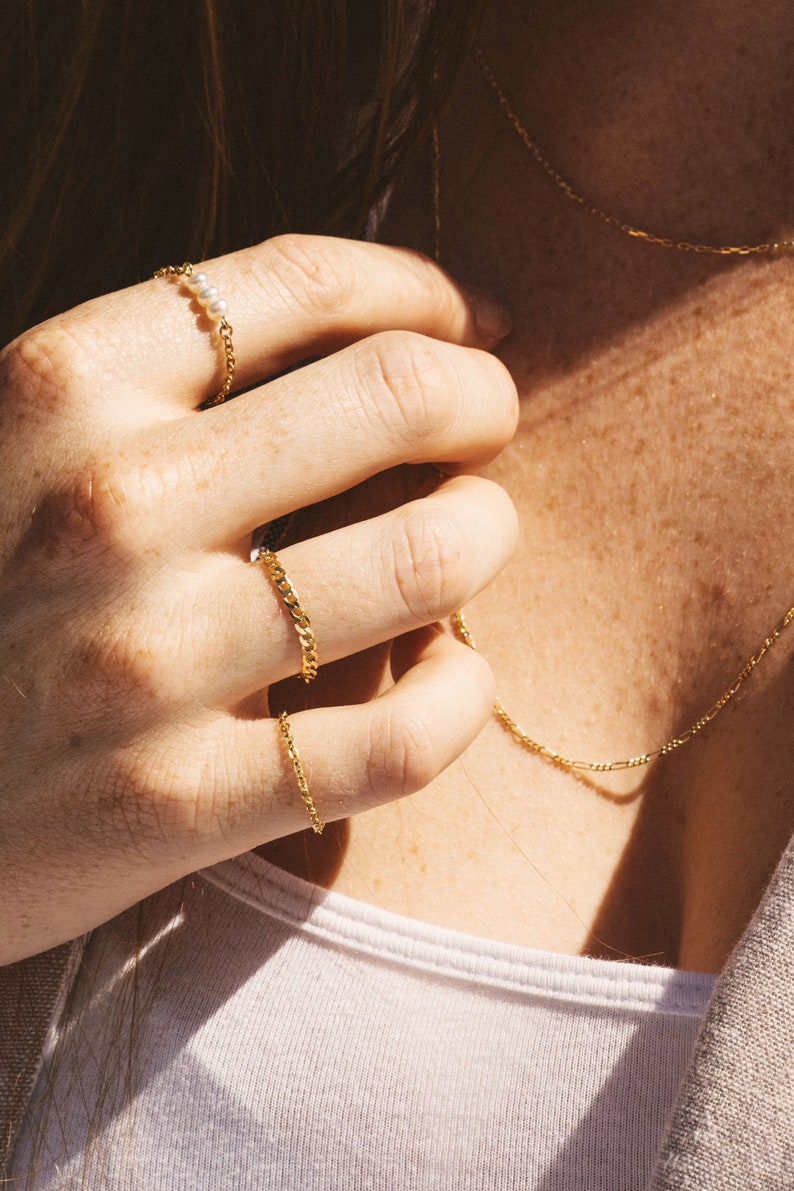 May include: A close-up shot of gold jewelry, including a chain necklace and three rings. One ring features a delicate chain design with three small pearls. The other two rings have a curb chain design. The jewelry is worn on a person's hand.
