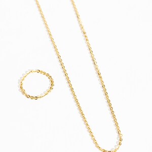 May include: A gold-colored necklace and ring set. The necklace is a delicate chain, and the ring is a matching band. Both pieces are displayed on a white background, creating a minimalist aesthetic. The jewelry appears to be made of metal.