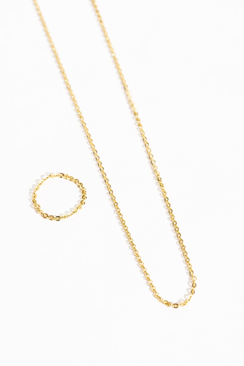 May include: A gold-colored necklace and ring set. The necklace is a delicate chain, and the ring is a matching band. Both pieces are displayed on a white background, creating a minimalist aesthetic. The jewelry appears to be made of metal.