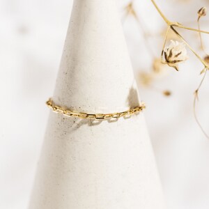 K&ouml;nnte beinhalten: Ein goldenes Kettenarmband mit einem zarten, quadratischen Gliederdesign. Das Armband wird auf einem wei&szlig;en, kegelf&ouml;rmigen St&auml;nder pr&auml;sentiert.